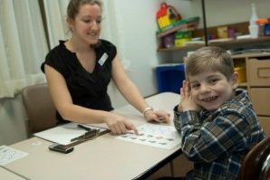A young boy smiles at the camera while he sits in a session with a female student clinician